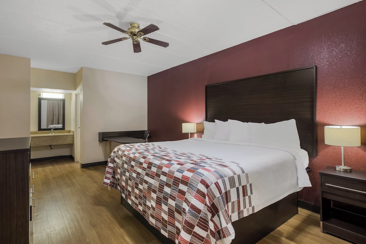 Een hotelkamer met een groot bed, een plafondventilator, een nachtkastje met lamp, een houten dressoir en een badkamer zichtbaar door een geopende deur.