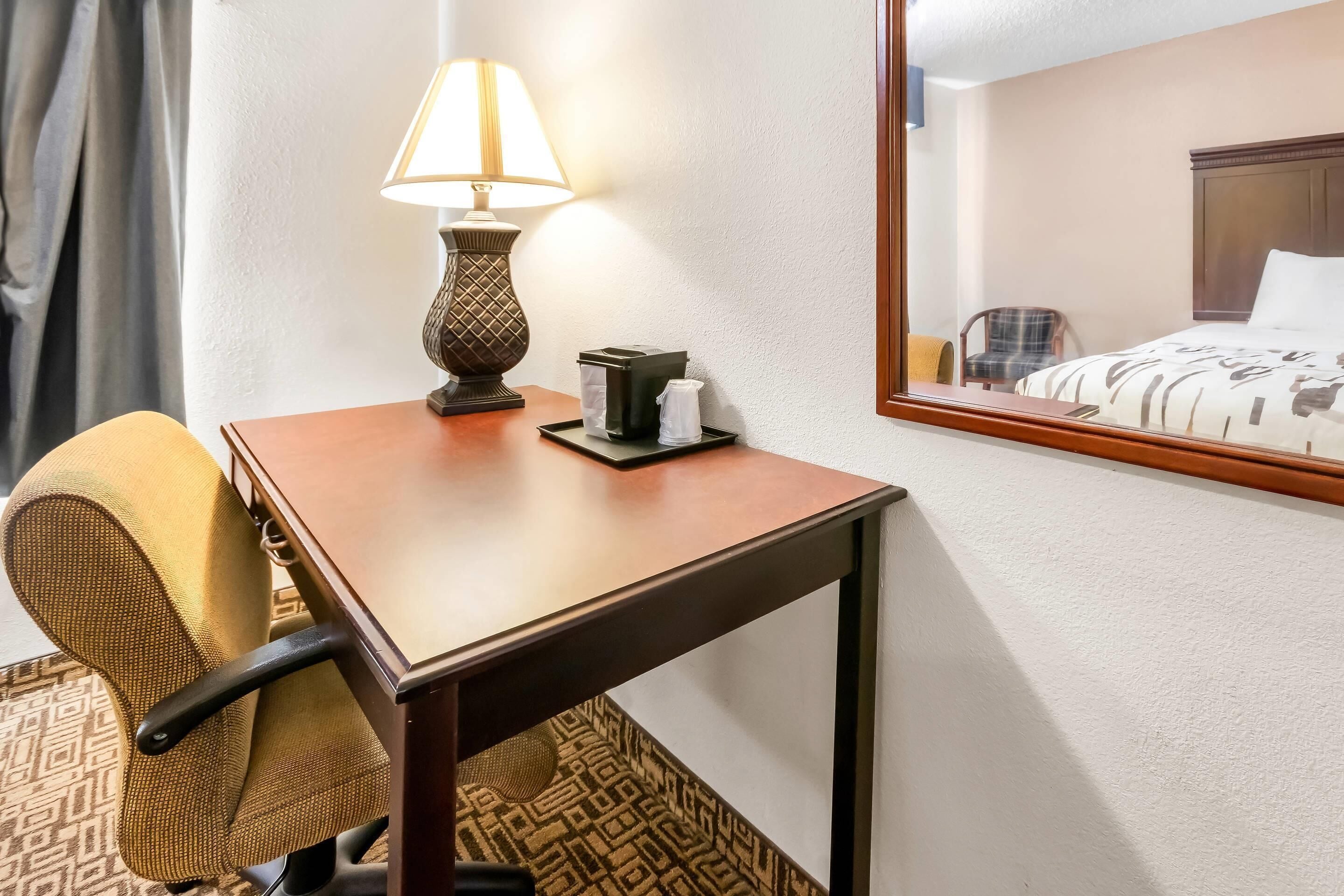 A hotel room with a wooden desk, a lamp, a chair, a bed with a patterned bedspread, and a mirror reflecting the room.