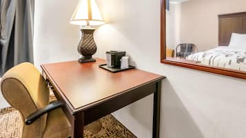 A hotel room with a wooden desk, a lamp, a chair, a bed with a patterned bedspread, and a mirror reflecting the room.