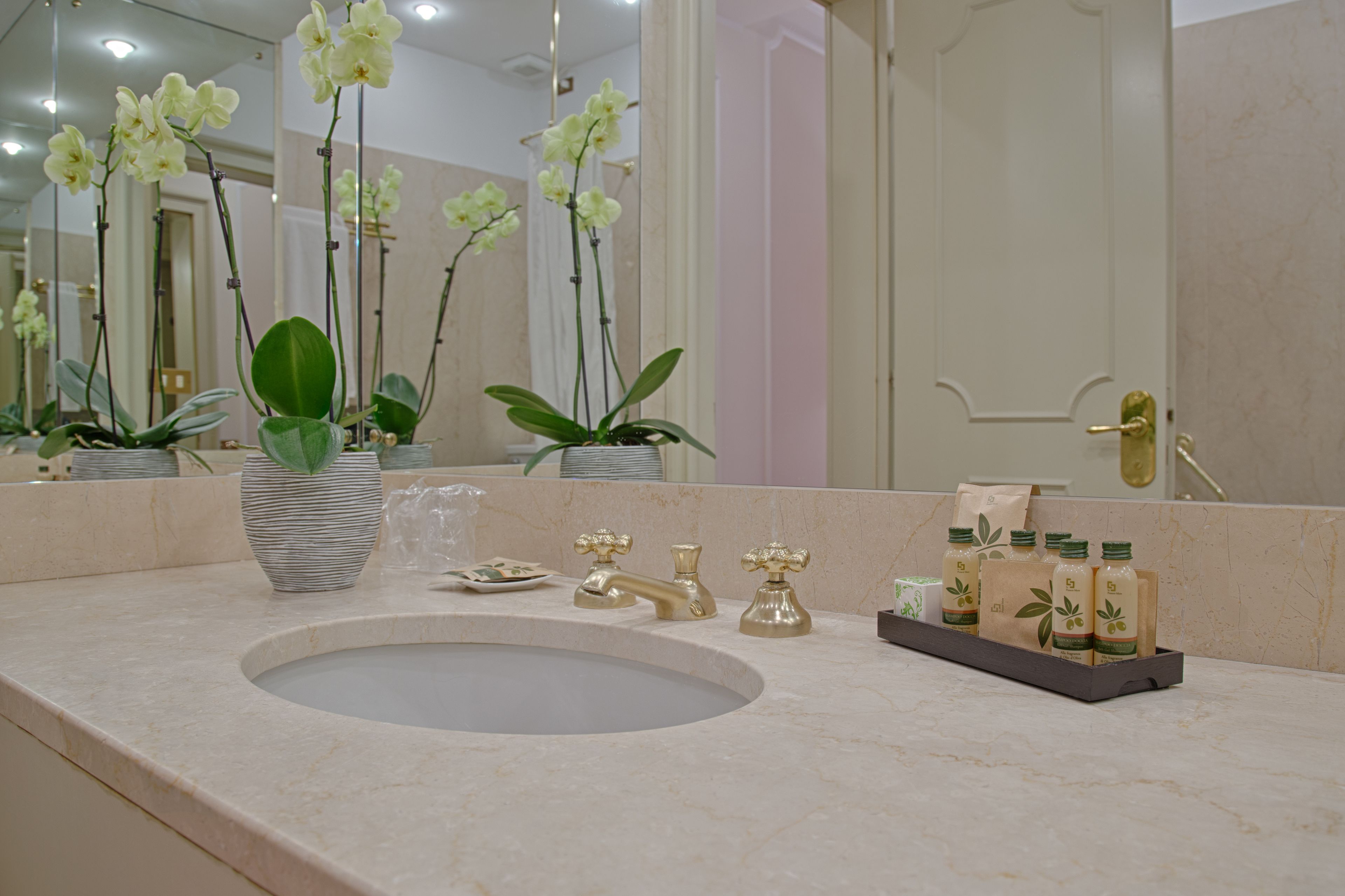 A bathroom with a large sink, two gold faucets, and orchid plants.