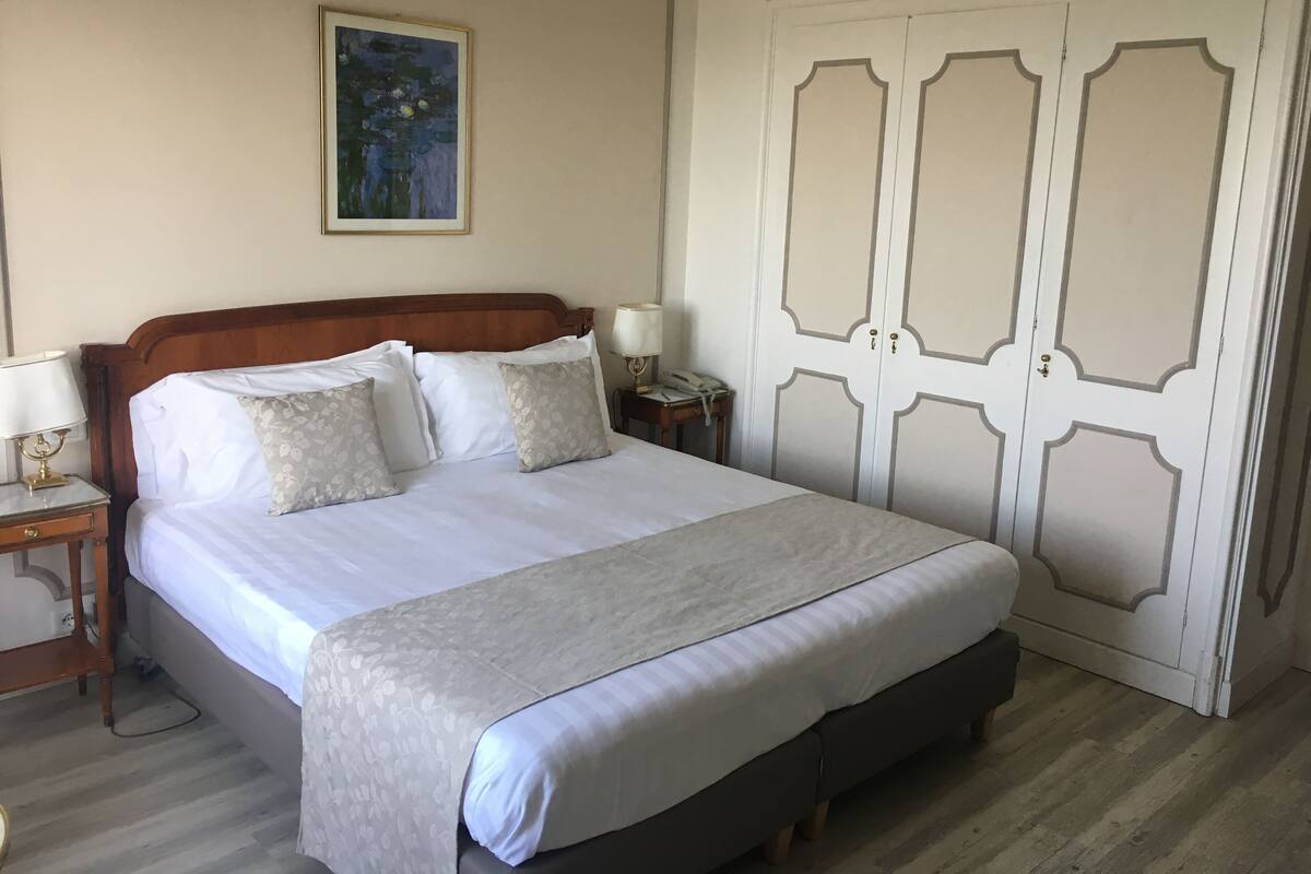 Un lit bien fait, avec du linge de lit blanc et des coussins à motifs floraux, une table de chevet avec une lampe et une armoire au décor élégant.