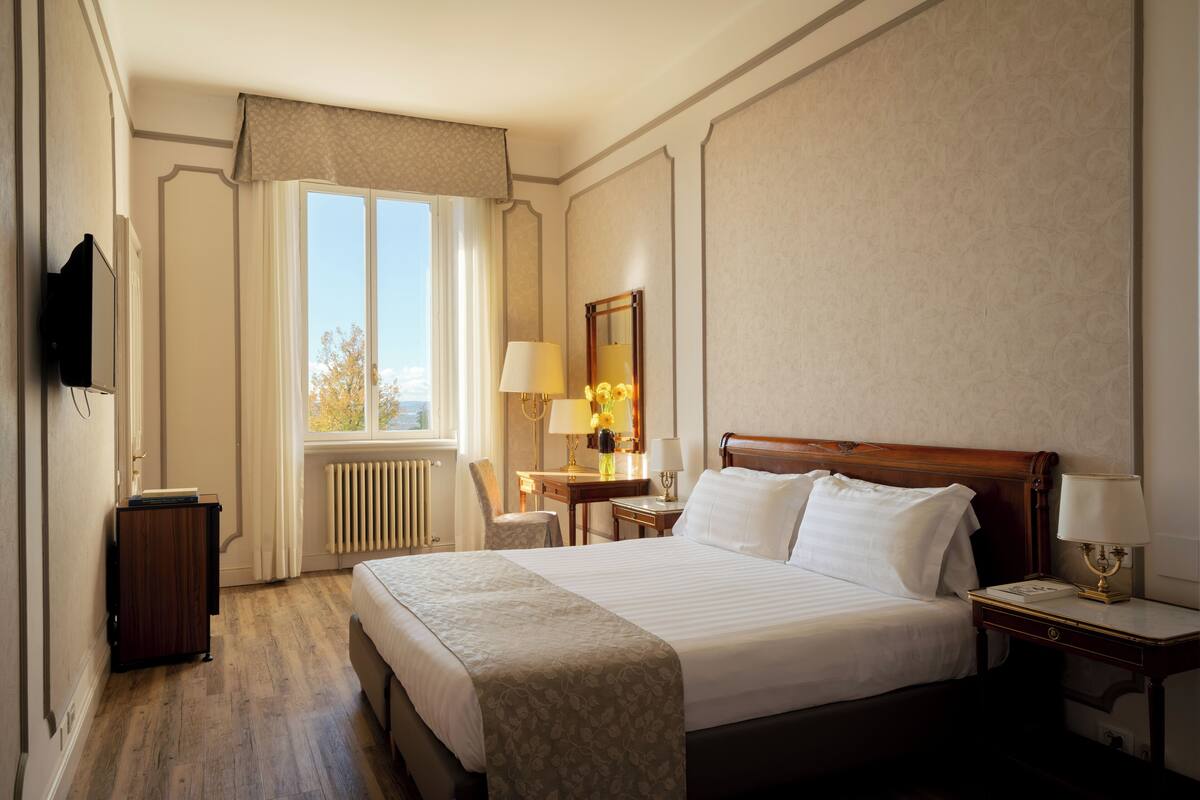 Une chambre d’hôtel avec un grand lit, un bureau avec une lampe, une chaise, une télévision et une fenêtre avec des rideaux.