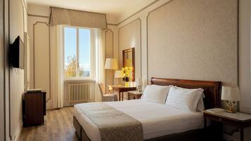 A hotel room with a large bed, a desk with a lamp, a chair, a television, and a window with curtains.