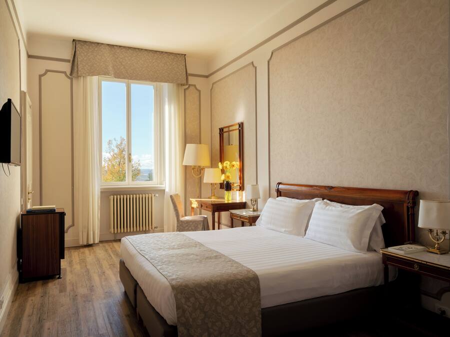 A hotel room with a large bed, a desk with a lamp, a chair, a television, and a window with curtains.