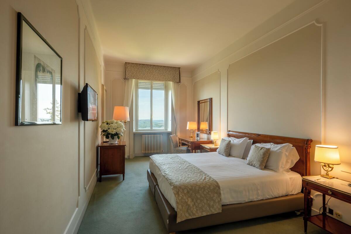 Une chambre d’hôtel avec un grand lit, des lampes de chevet, un bureau avec une chaise, un miroir et une fenêtre avec des rideaux.