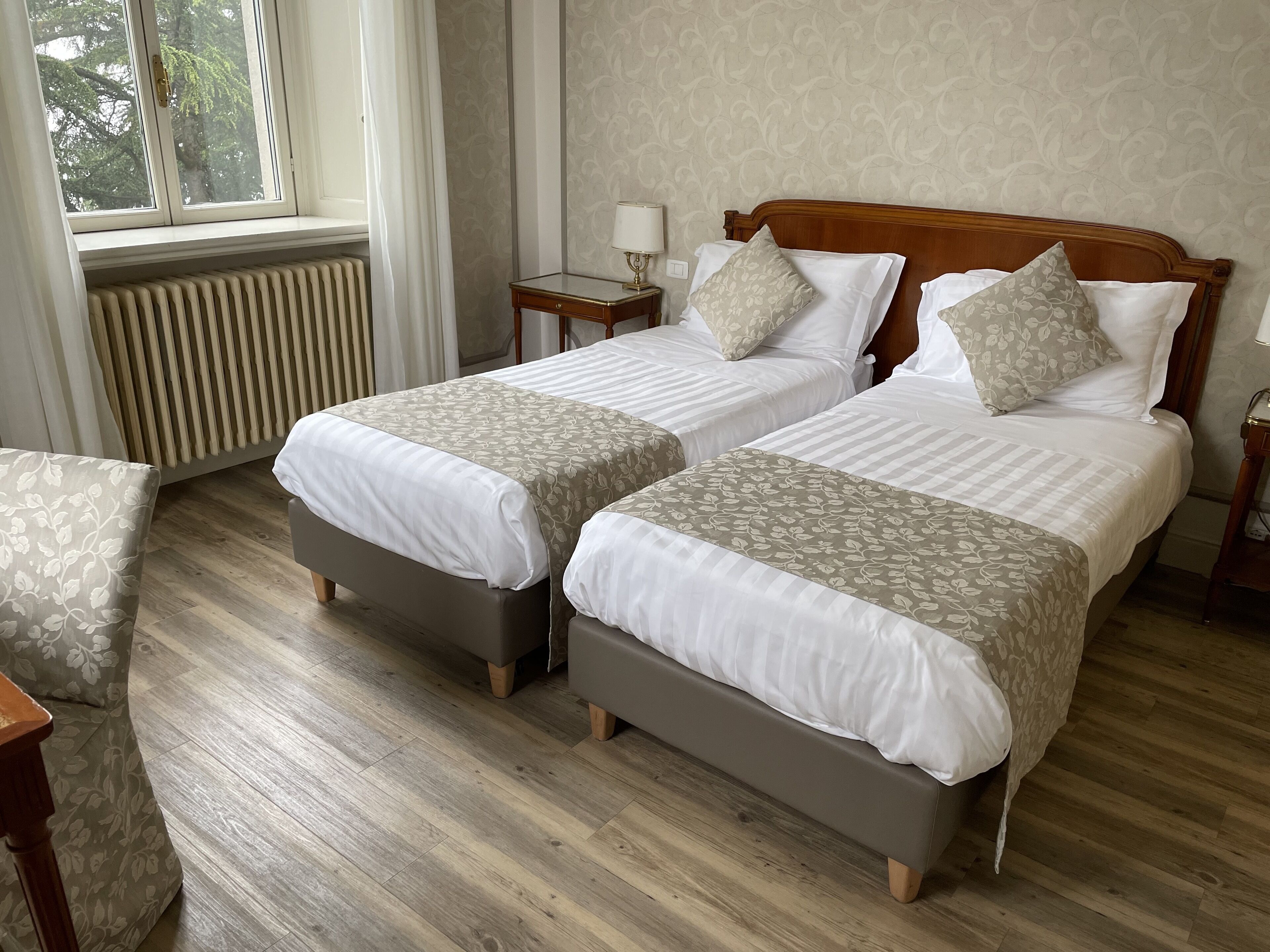 A hotel room with two beds, a radiator, a window with curtains, and a wooden floor.
