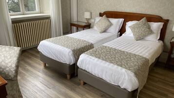 A hotel room with two beds, a radiator, a window with curtains, and a wooden floor.