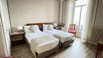 Habitación de hotel con dos camas, una silla, una mesita de noche con lámpara y una ventana con cortinas.