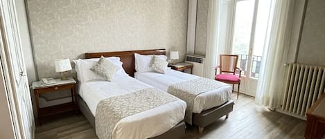 A hotel room with two beds, a chair, a nightstand with a lamp, and a window with curtains.