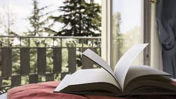 An open book on a bed with a view of a wooden deck and trees outside a window.
