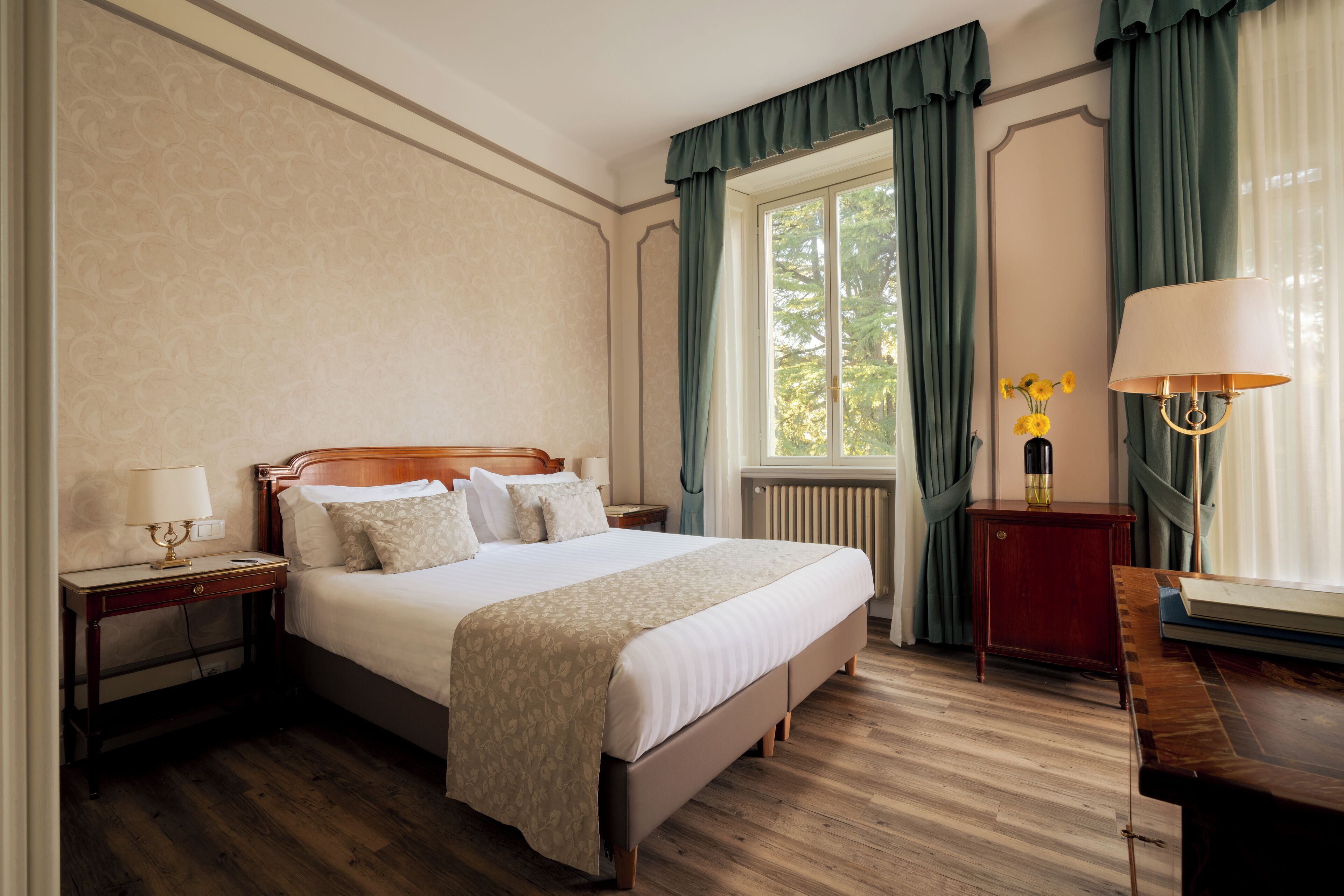 Ein ordentlich eingerichtetes Schlafzimmer mit einem großen Bett, einem Nachttisch mit Lampe, einem kleinen Tisch mit einer Vase und Blick ins Grüne durch das Fenster.