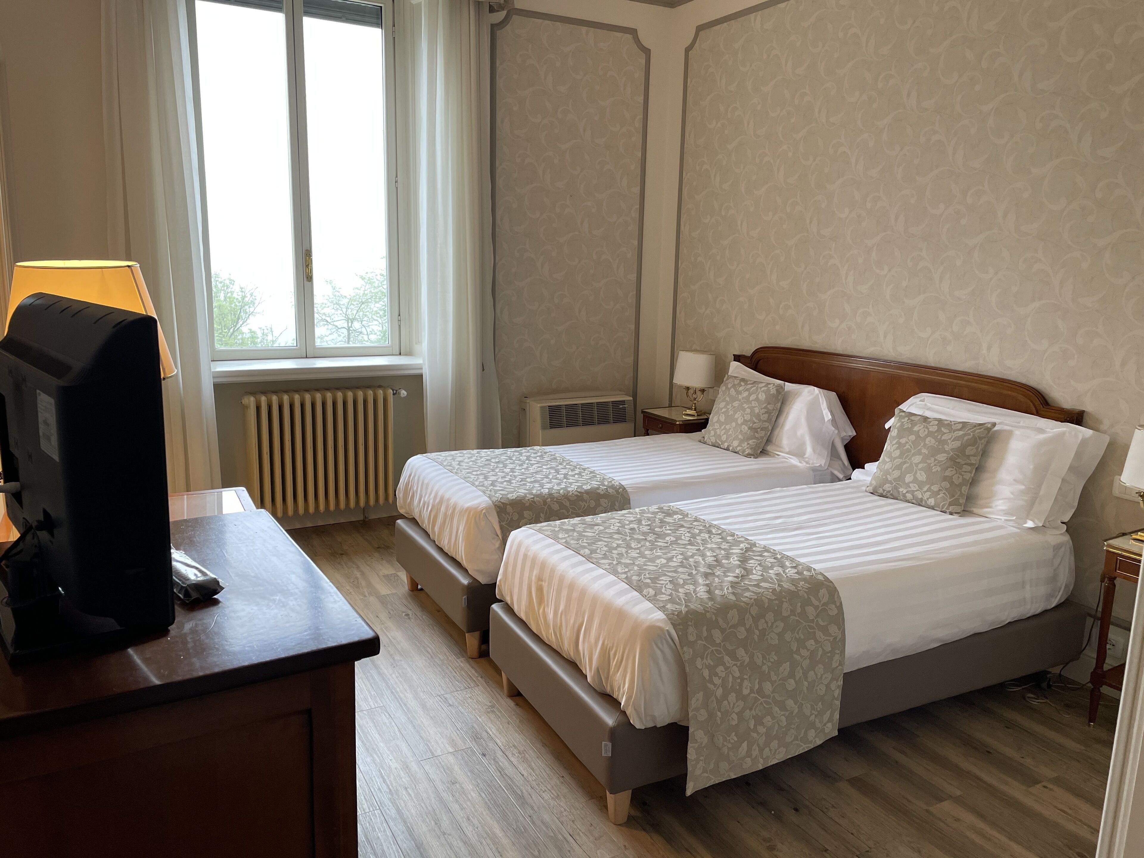 A hotel room with two beds, a desk with a lamp, a radiator, and a window with curtains.