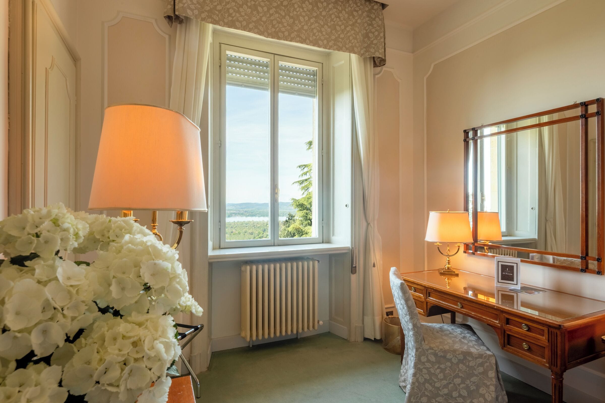 A hotel room with a large window, a wooden desk with two lamps, a mirror, and a floral arrangement.
