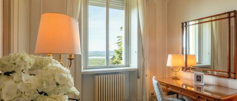 Ein Hotelzimmer mit großem Fenster, einem Holztisch mit zwei Lampen, einem Spiegel und einer Blumenkomposition.
