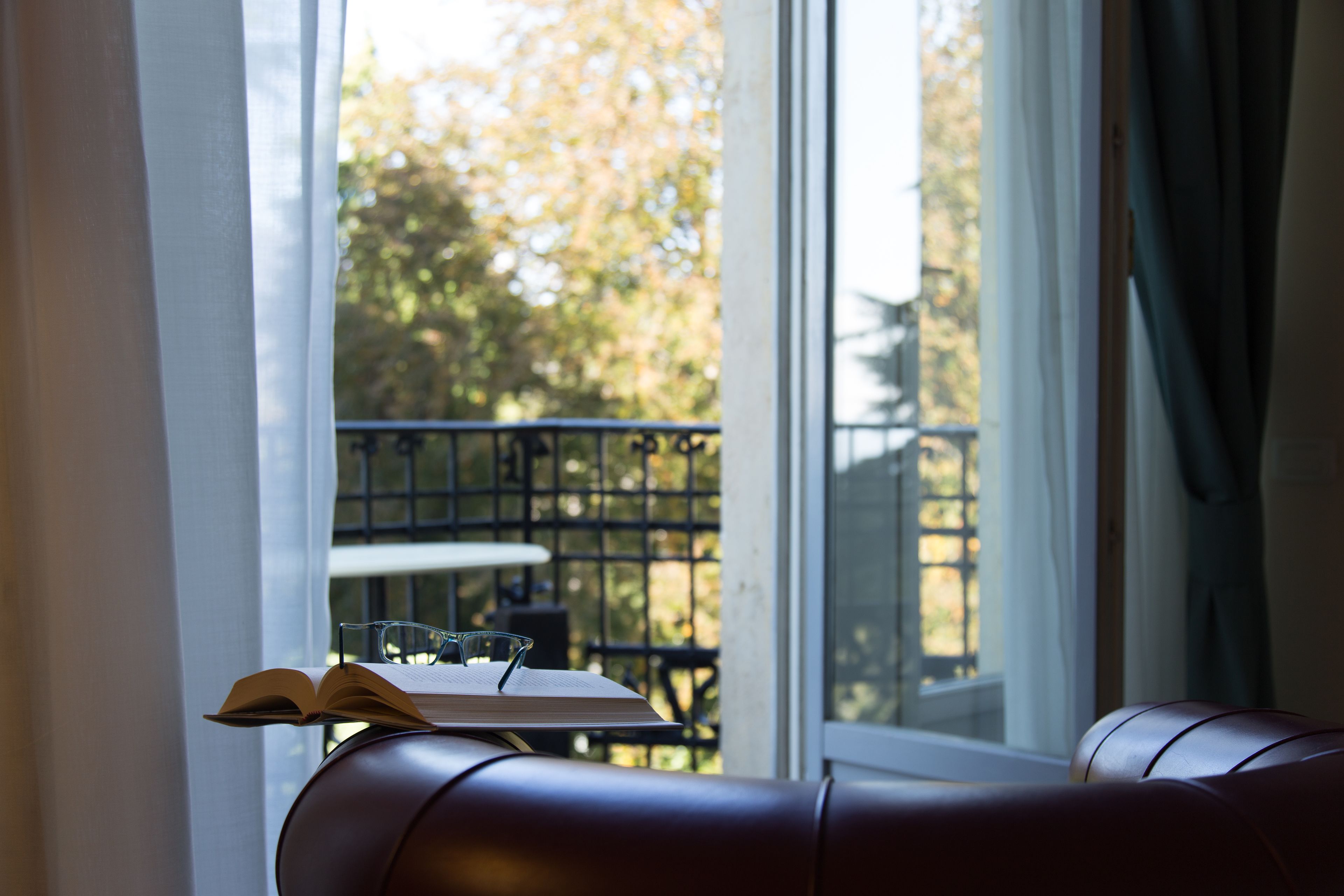 A leather couch with a book and glasses, a window with a view of a balcony and trees.