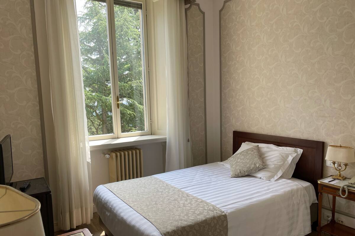 Un lit bien fait, recouvert d’une couvre-lit à motifs floraux, une table de chevet avec une lampe et une fenêtre avec des rideaux.