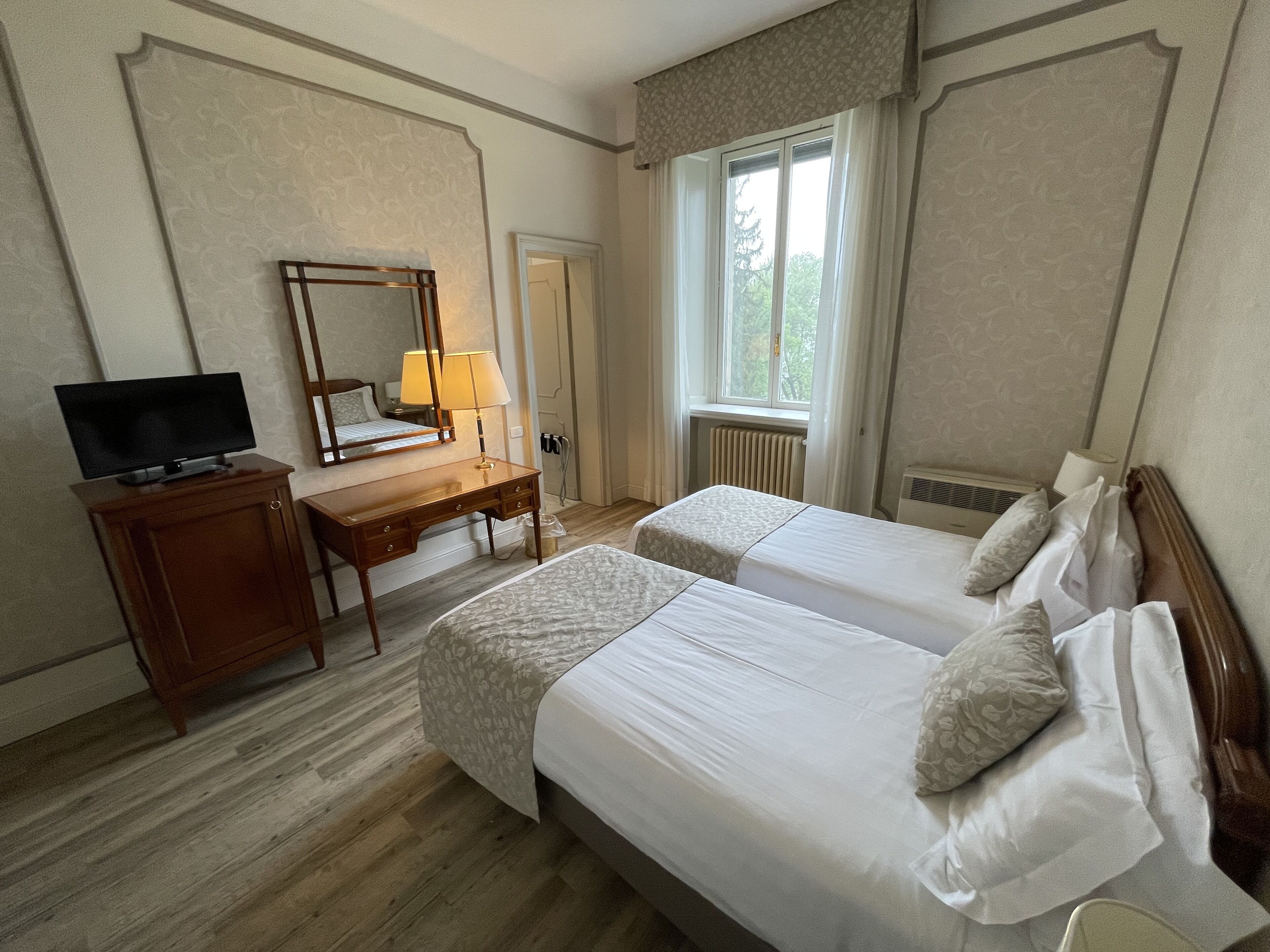 Habitación de hotel con dos camas, un escritorio con televisor, un espejo y ventana con vista a árboles.
