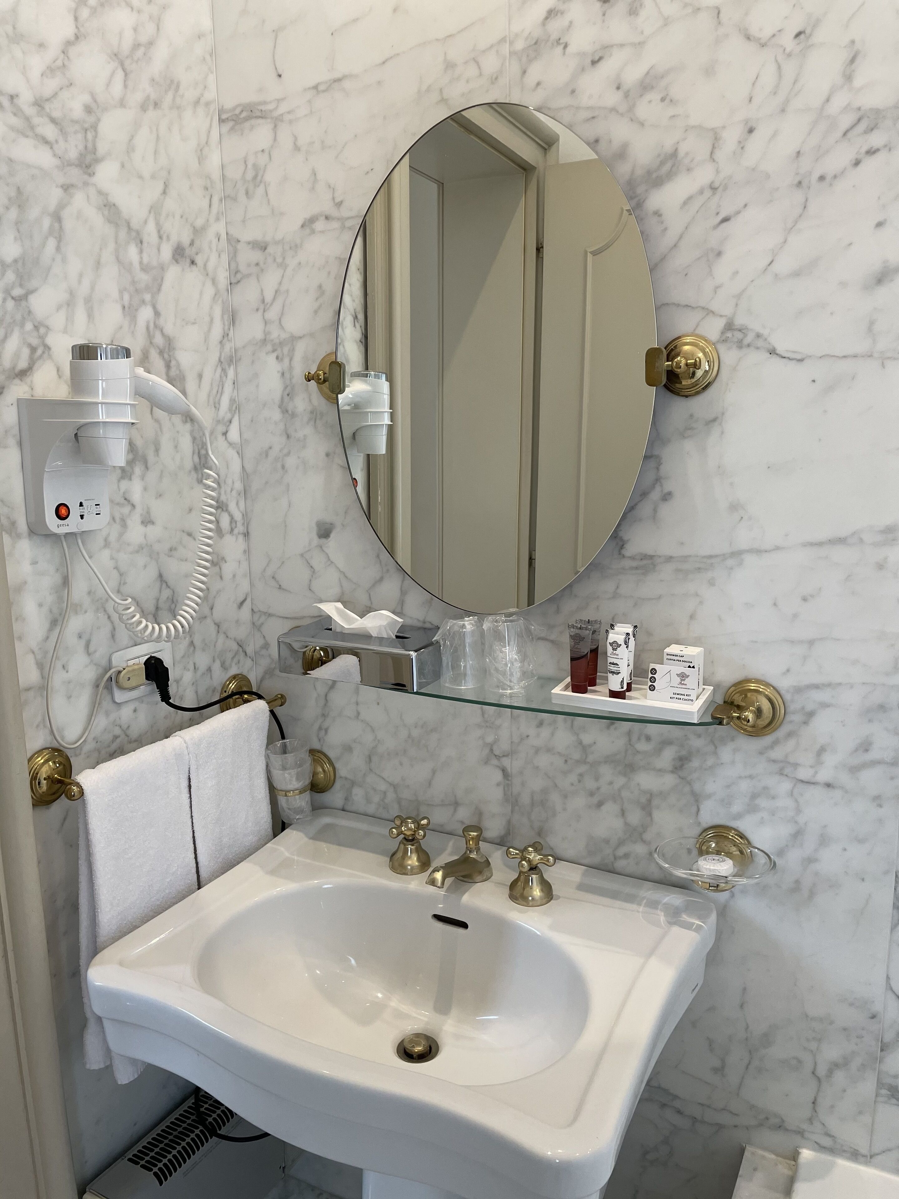 A bathroom with a marble wall, a round mirror, a white sink with gold faucets, and a towel rack.