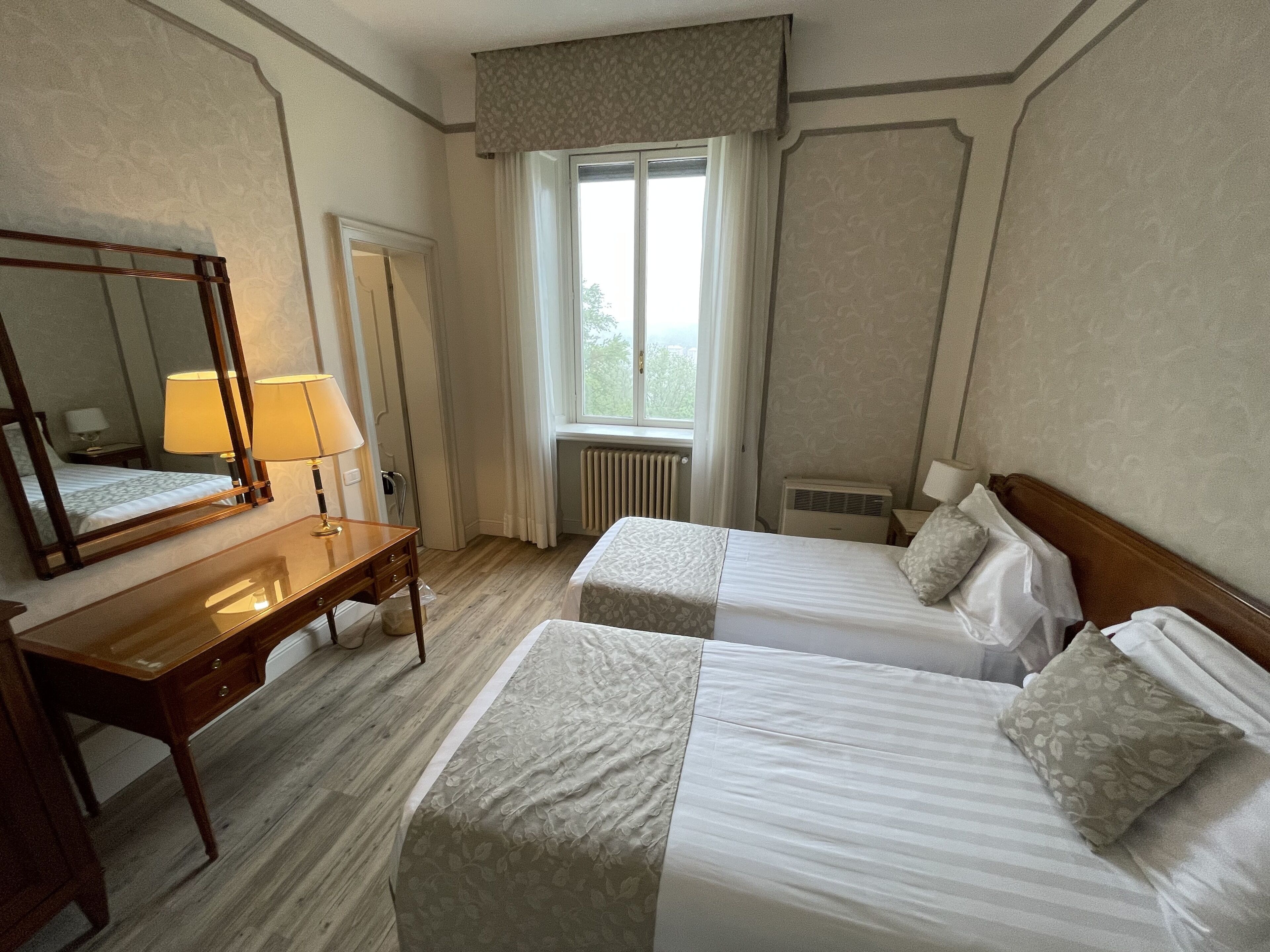 Une chambre d’hôtel avec deux lits simples, un bureau en bois avec une lampe, un grand miroir et une fenêtre avec des rideaux.