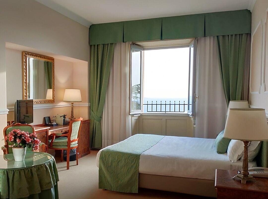 A hotel room with a bed, a desk with a chair, a table with a vase of flowers, a lamp, and a window with curtains.