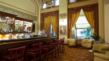 A bar area with multiple liquor bottles, bar stools, and a chandelier. There are two windows with curtains, a painting on the wall, and a potted plant near the windows.