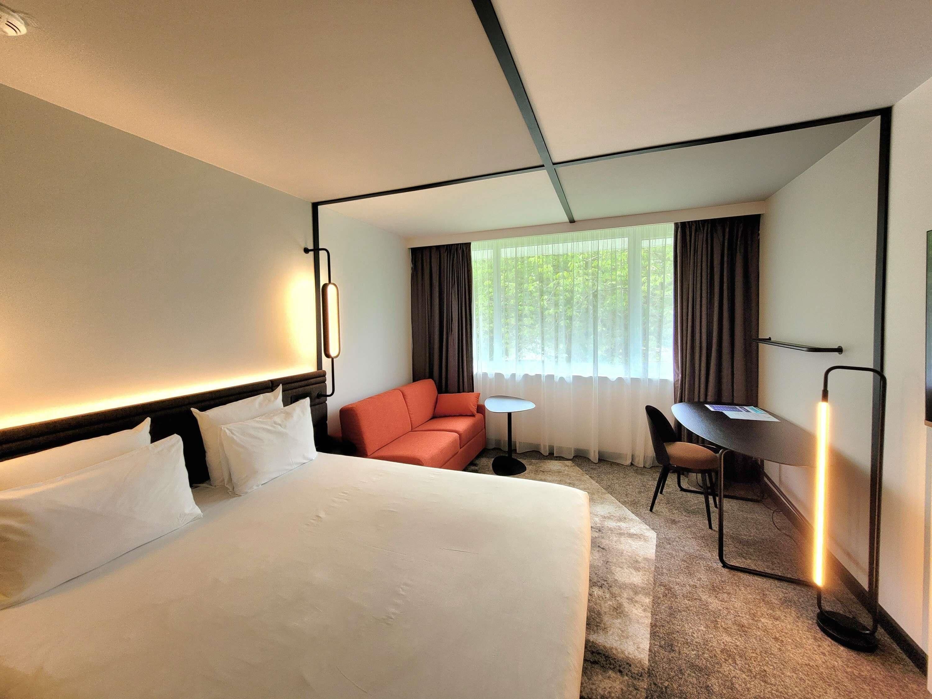 A hotel room with a bed, a desk, a chair, a sofa, and a window with curtains.