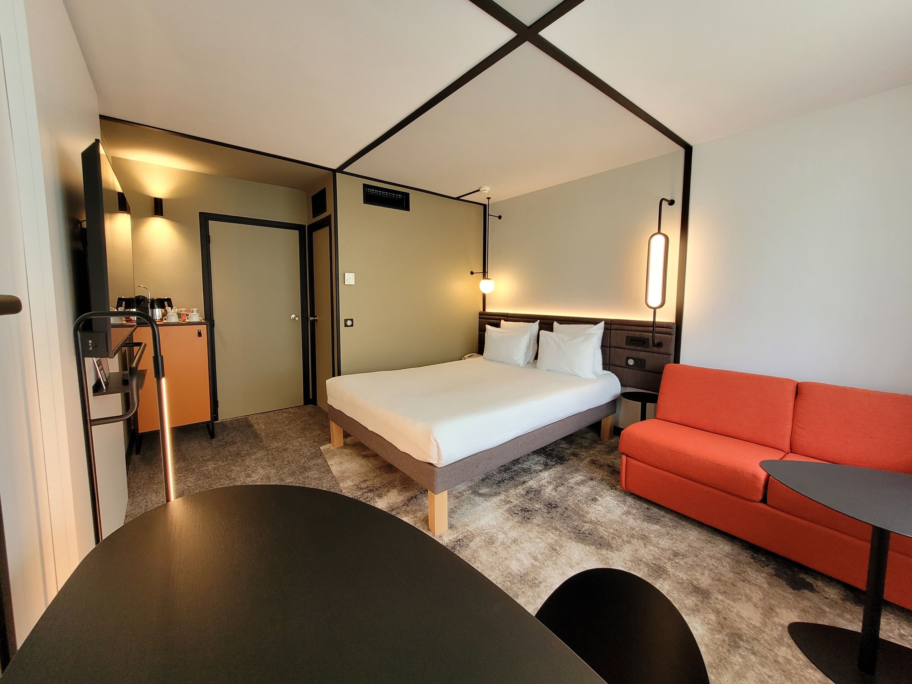 Habitación de hotel con una cama, un sofá rojo, una mesita, un escritorio con una silla y una lámpara de pared.