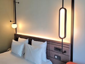 A hotel room with a neatly made bed, a bedside table, a wall-mounted light fixture, and a mirror. - Novotel Lille Aeroport (Lesquin)