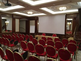 Μια αίθουσα συνεδριάσεων με σειρές από κόκκινες καρέκλες, μια λευκή οθόνη προβολής και πολυελαίους.