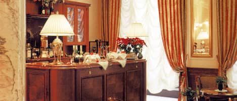 A hotel lobby with a marble column, a reception desk, a dining area with a table and chairs, and a large window with curtains.