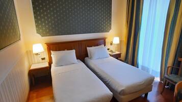 Una habitación de hotel con dos camas individuales, cabeceros de madera, mesitas de noche con lámparas, un espejo grande y vistas al exterior a través de cortinas.
