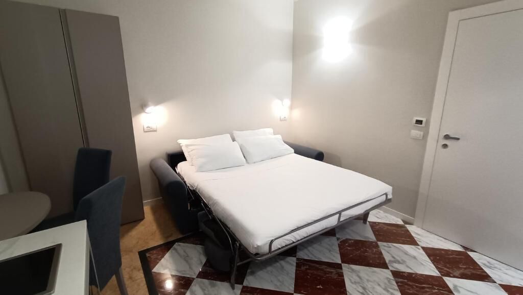 A small, neatly arranged room with a bed, bedside table, chair, and a small desk with a chair.