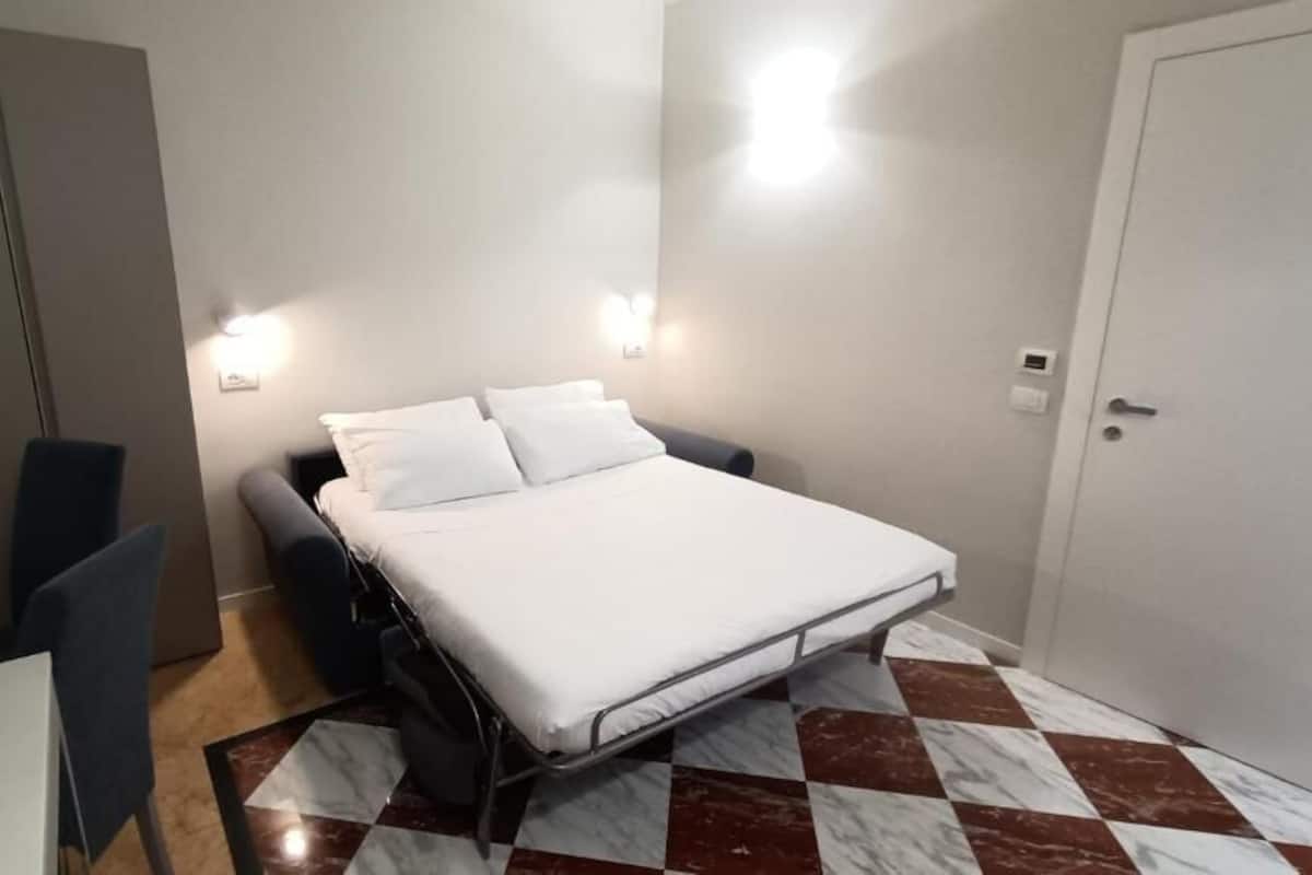 Una habitación pequeña y ordenada con cama, mesita de noche, silla y un escritorio con silla.