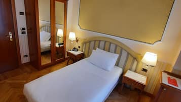 A hotel room with a single bed, wooden floor, a wardrobe, a bedside table with a lamp, and a large mirror.