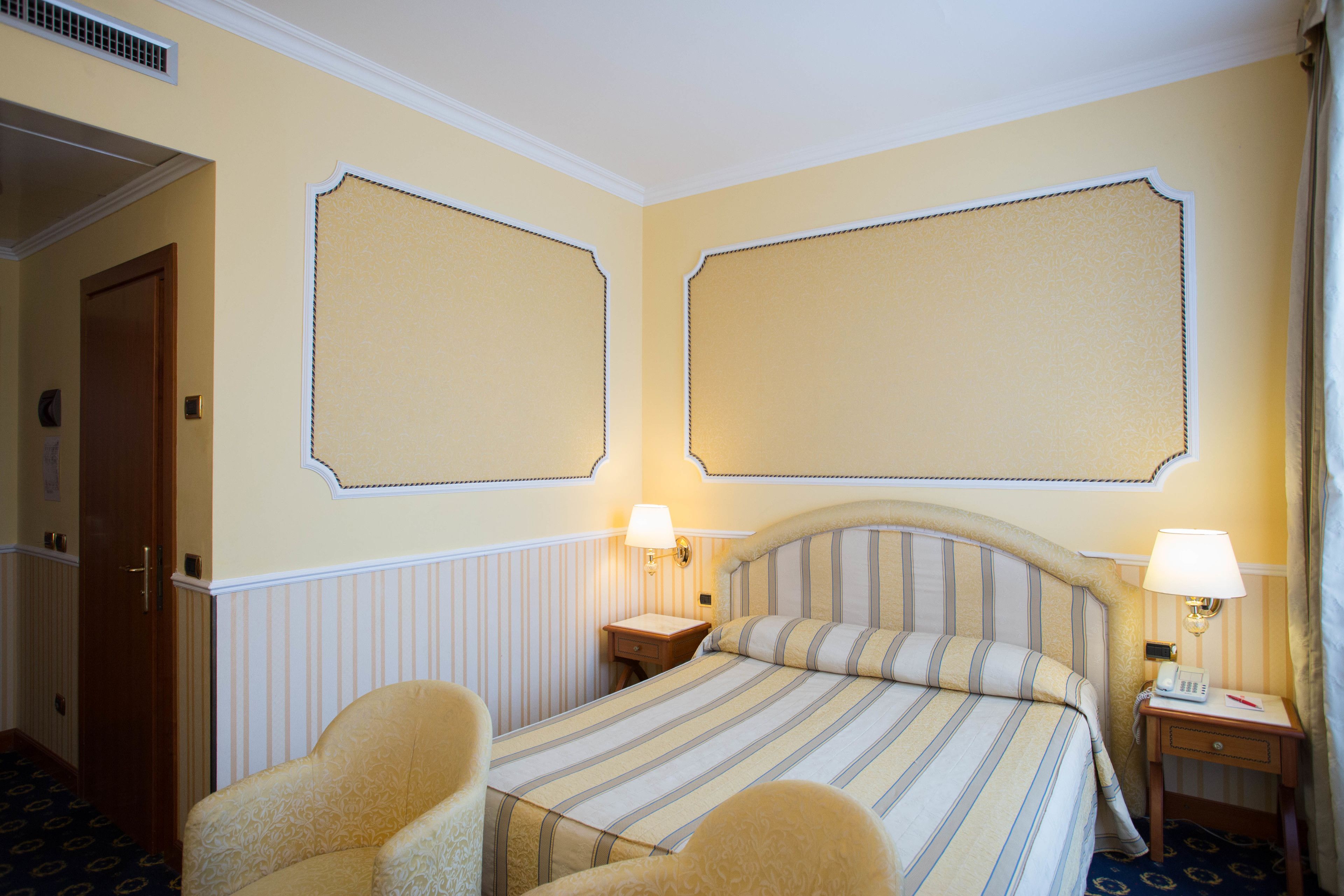 Quarto de hotel com uma cama grande, duas mesas de cabeceira com abajures, uma porta de madeira e um tapete estampado.