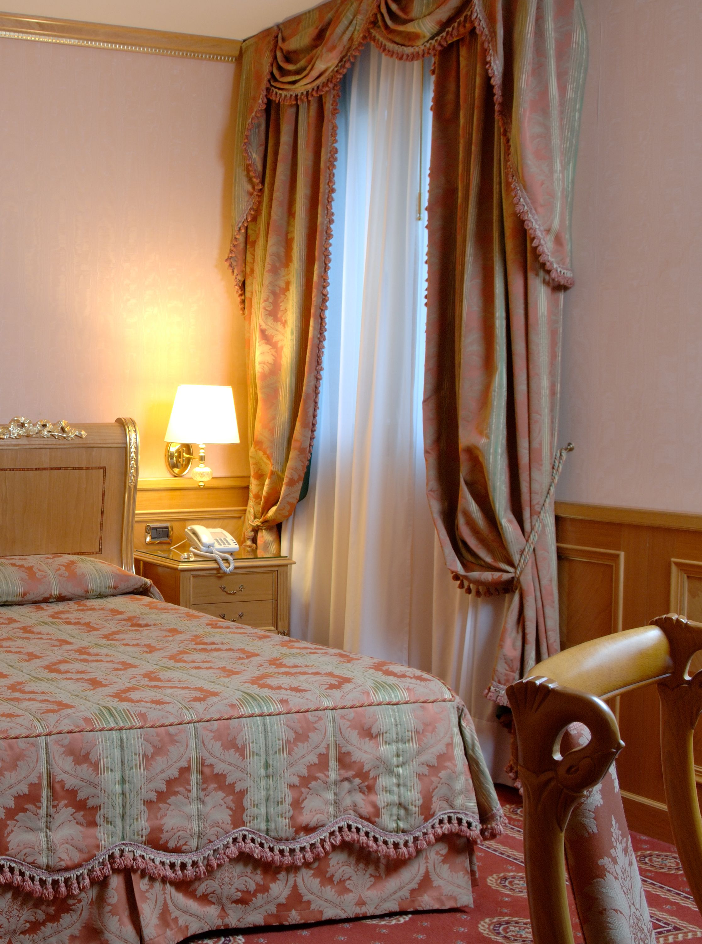Quarto de hotel com cama, mesa de cabeceira com abajur, telefone e uma cadeira.