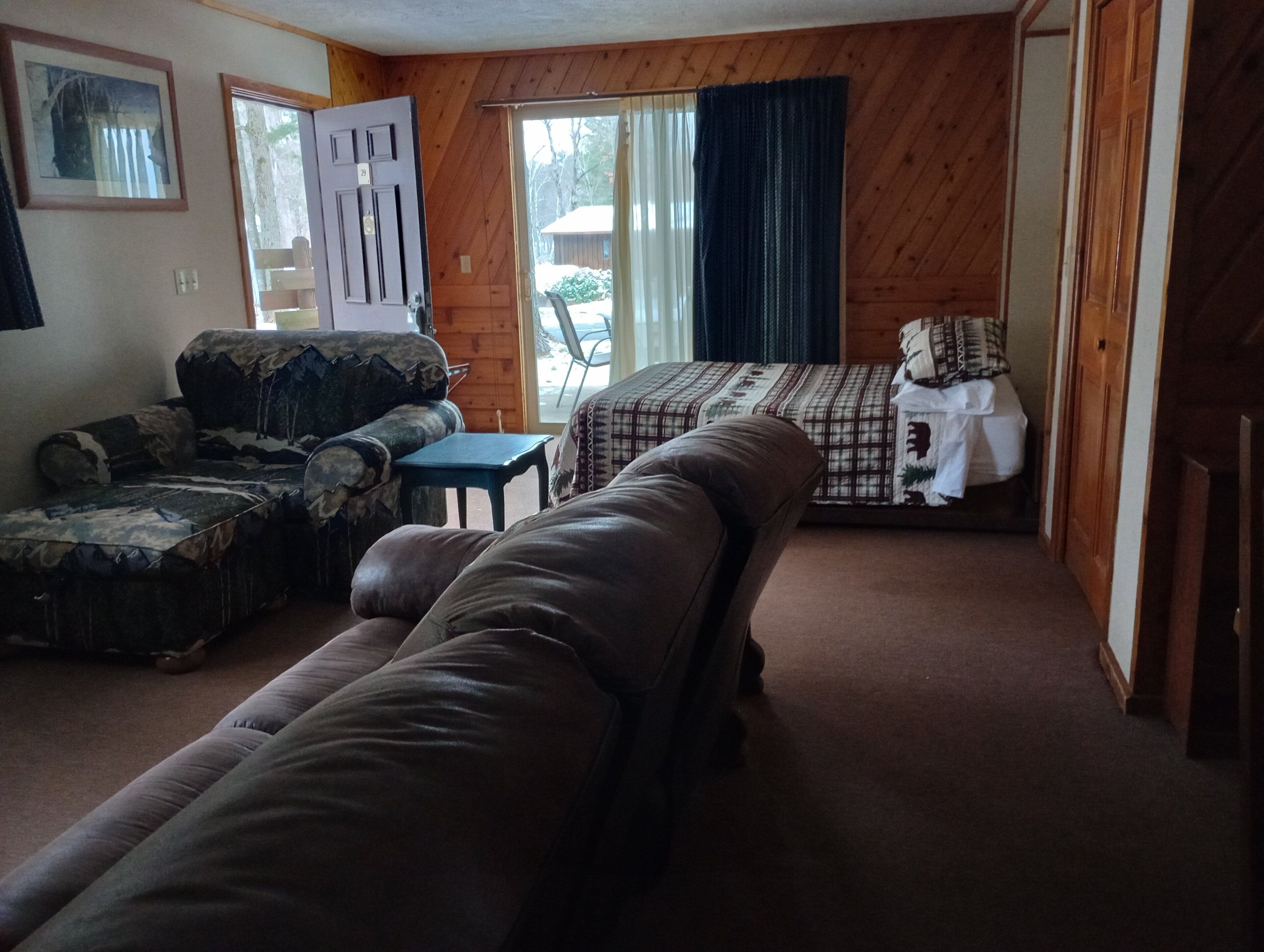 A room with a brown leather sofa, a patterned armchair, a small table, and a bed with a checkered bedspread.