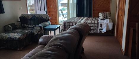 A room with a brown leather sofa, a patterned armchair, a small table, and a bed with a checkered bedspread.