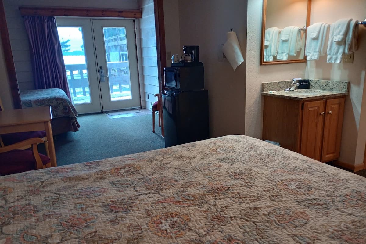 A hotel room with a large bed, a desk, a chair, a kitchenette, and a bathroom with towels and a mirror.