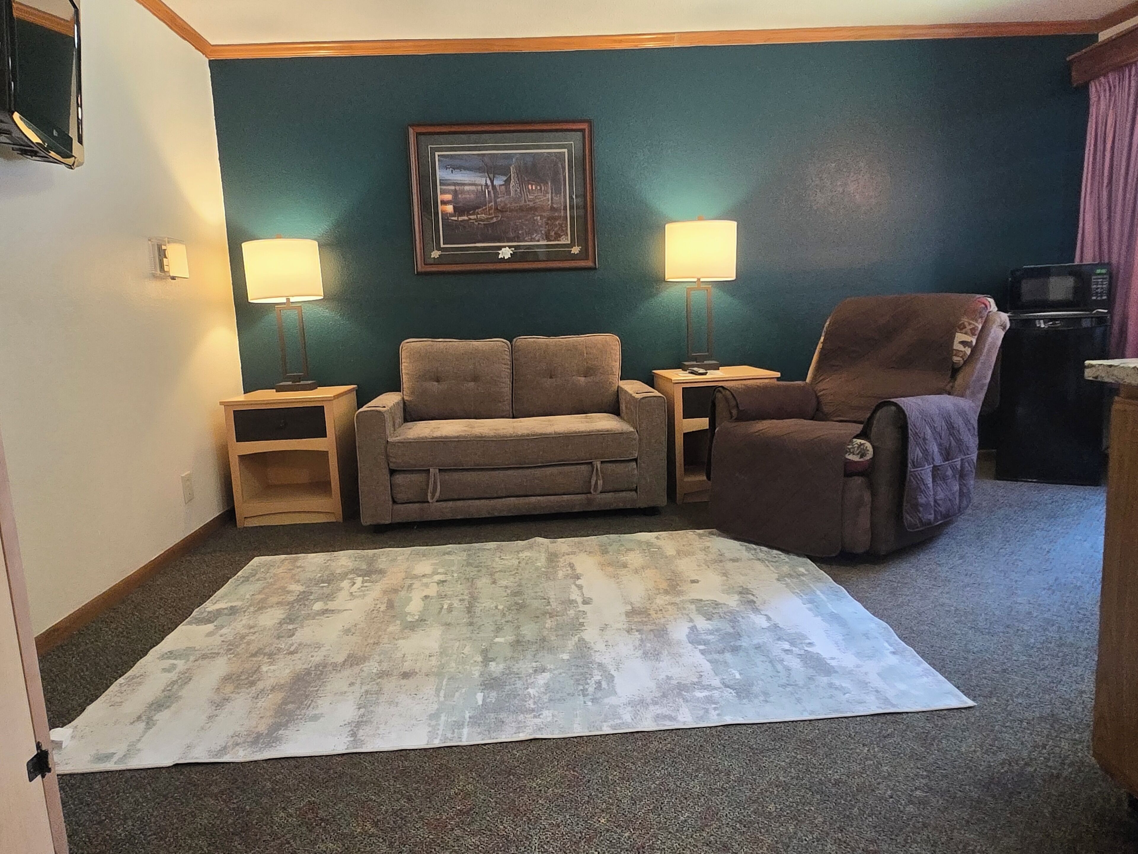 Una sala de estar con sofá, sillón, dos mesitas auxiliares, lámpara, un cuadro enmarcado y televisor.