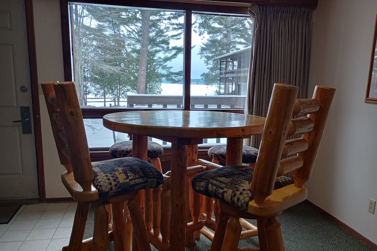 A wooden dining table with four chairs, a view of a deck and trees through a large window, and a door with a doorknob.