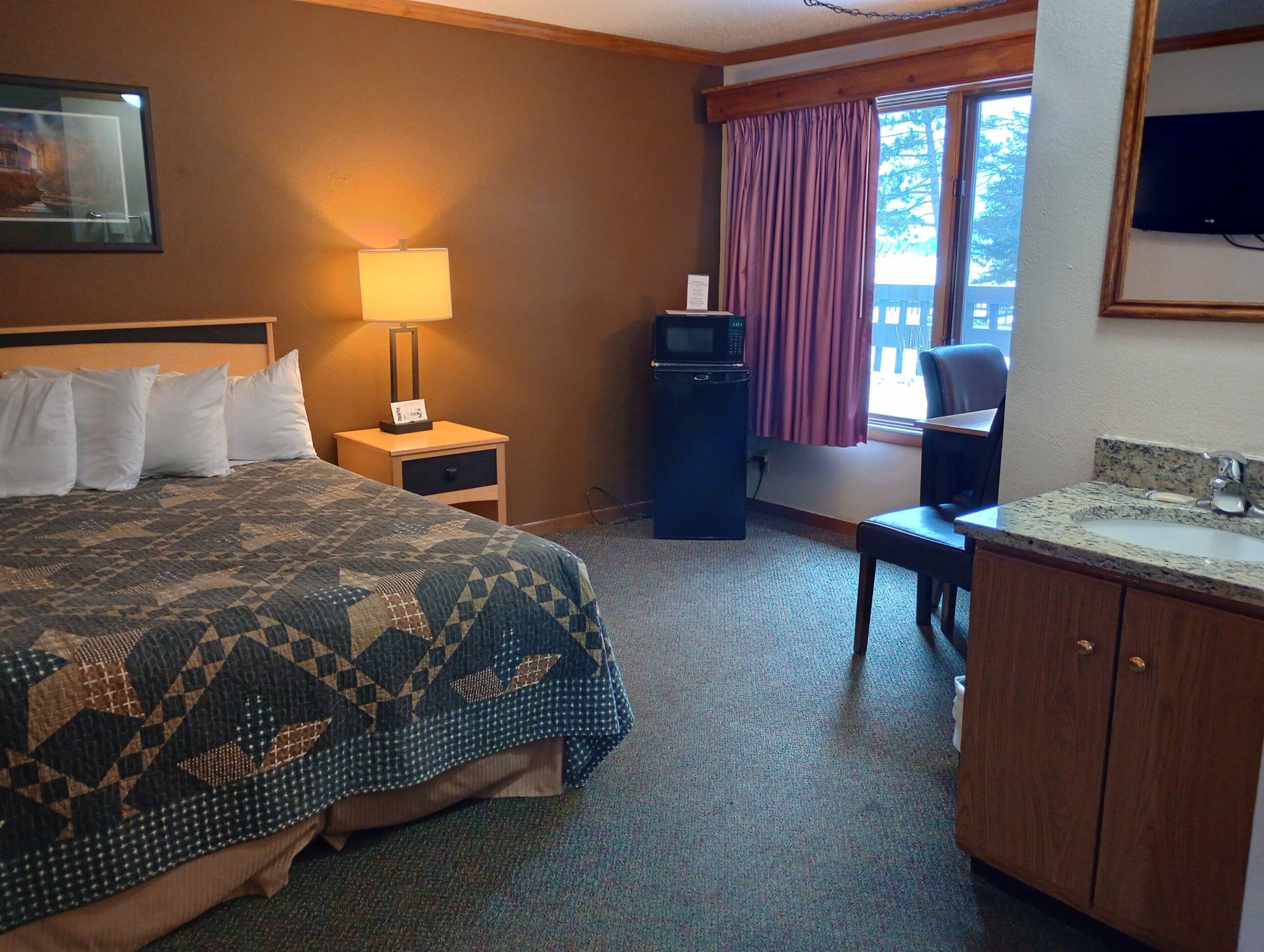 A hotel room with a bed, a desk, a television, a window with curtains, and a sink.