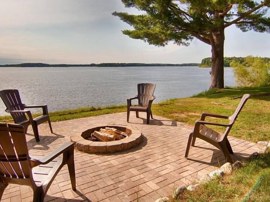 Eine Terrasse mit vier Adirondack-Stühlen, einer runden Feuerstelle und Blick auf einen See.