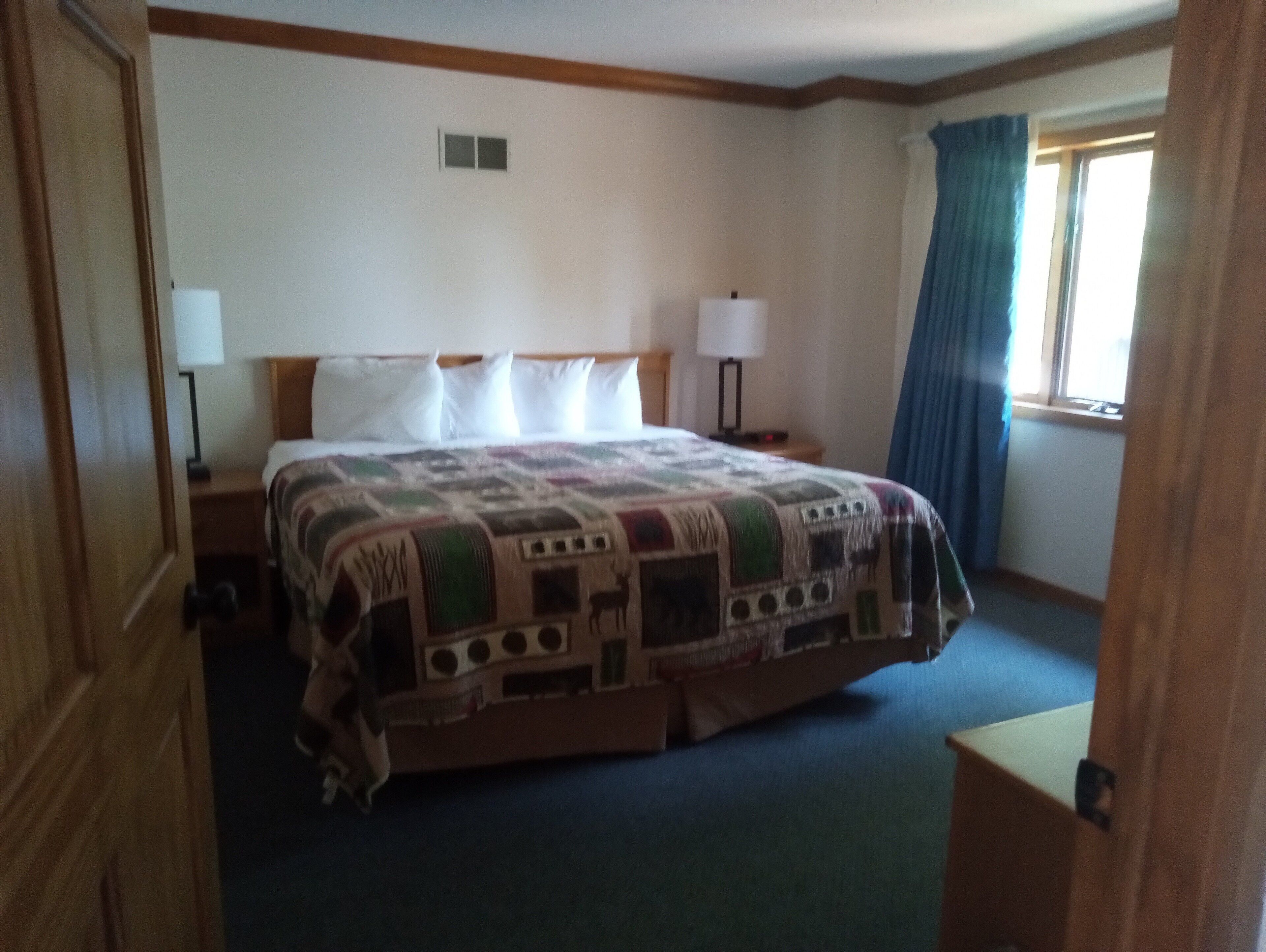 Habitación de hotel con una cama grande, dos mesitas de noche, una ventana con cortinas y una puerta.