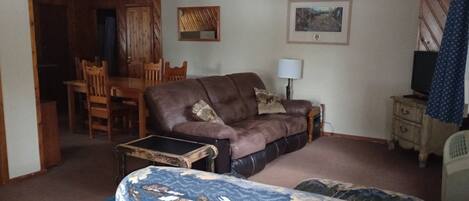 A living room with a brown leather sofa, a blue armchair, a wooden dining table with chairs, a small cabinet, and a television on a wooden stand.