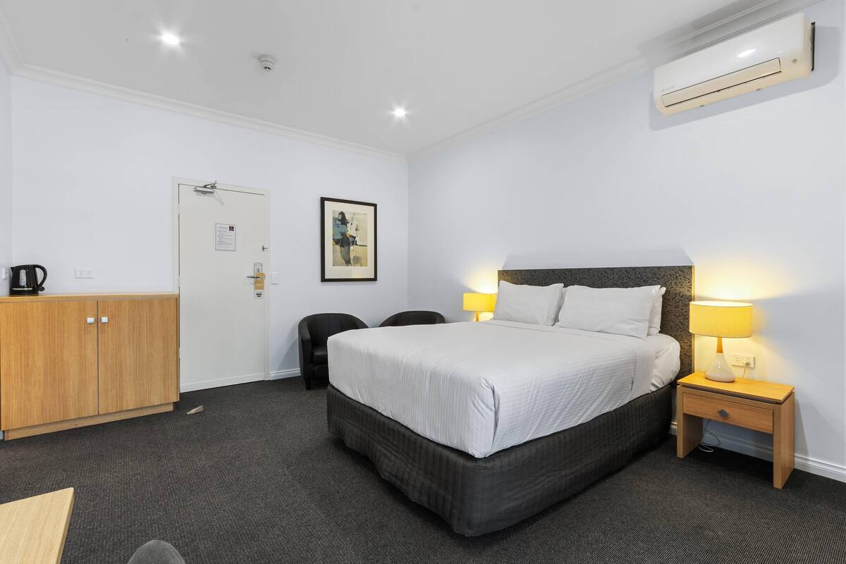 Quarto de hotel com cama, mesa de cabeceira, luminária, ar condicionado, guarda-roupa e porta com uma placa.