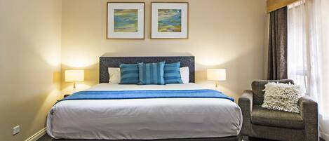 Habitación de hotel con una cama grande, dos lámparas de noche, un sillón con almohadón y dos cuadros en la pared.