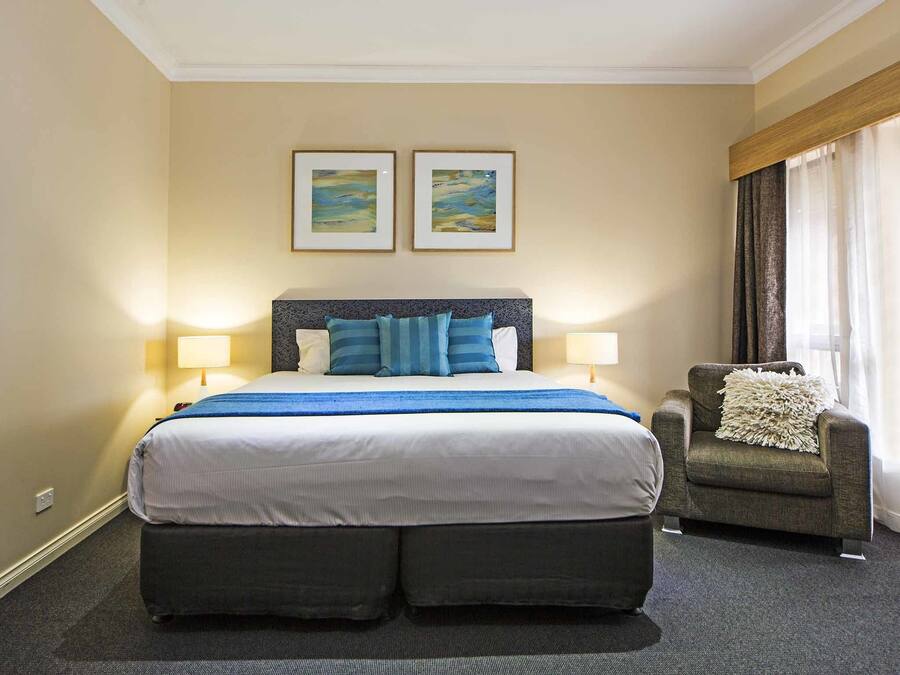 A hotel room with a large bed, two bedside lamps, a chair with a throw pillow, and two framed pictures on the wall.