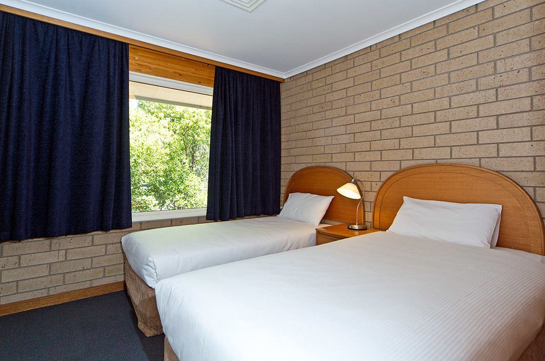 A hotel room with two single beds, a wooden headboard, a nightstand with a lamp, and a brick wall.