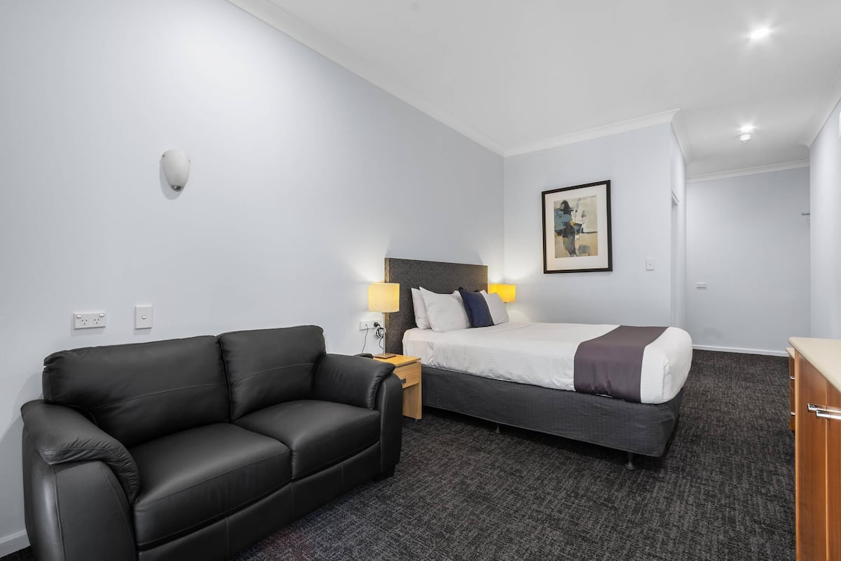 Quarto de hotel com sofá de couro preto, cama com roupa de cama branca e escura, quadro na parede e mesa de cabeceira de madeira pequena com abajur.
